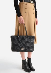 Borsa tote nera trapuntata con motivo intrecciato, dettagli in metallo dorato e tracolle in pelle. Abbinata a una gonna color cammello e stivaletti neri alla caviglia.