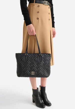 Borsa tote nera trapuntata con motivo intrecciato, dettagli in metallo dorato e tracolle in pelle. Abbinata a una gonna color cammello e stivaletti neri alla caviglia.