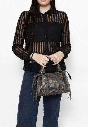 Piccola borsa in pelle grigia con doppi manici, dettagli metallici e frange. Il modello indossa una blusa a righe nere e jeans.