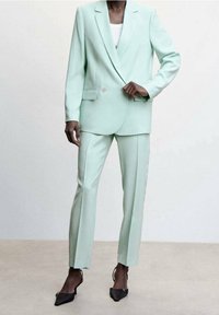 Femme portant un costume sur mesure vert menthe clair avec un blazer à un bouton et un pantalon droit, assorti à des escarpins noirs pointus.