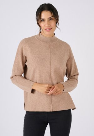 Mujer con cabello oscuro recogido, vistiendo un suéter de manga larga beige y pantalones negros, de pie frente a un fondo claro y liso.