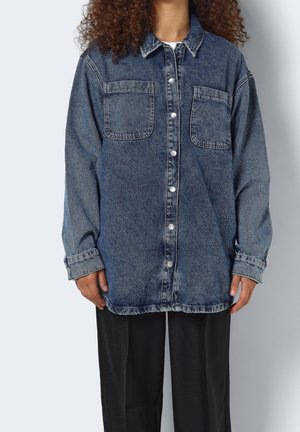 Personne portant une chemise en jean bleu foncé ample à boutons avec deux poches poitrine et un pantalon noir, debout devant un fond clair.