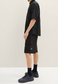 Muž v černé košili s krátkým rukávem na knoflíky, černých cargo šortkách s kapsou na klopě, černých ponožkách a černých nízkých teniskách, stojící bokem.