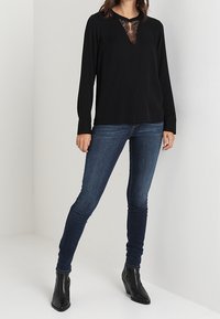 Top lungo nero a maniche lunghe con scollatura a V e inserti in pizzo, abbinato a jeans slim-fit blu scuro e stivaletti neri.