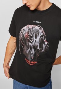 Schwarzes Baumwoll-T-Shirt mit einem grafischen Seitenschnitt, der strukturierte Designs zeigt, und roten Buchstaben, die "FLYING HIGH" am unteren Rand lesen.