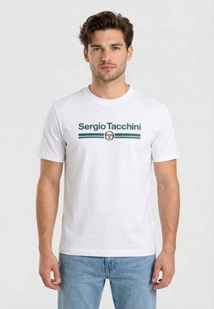 Junger Mann mit kurzen braunen Haaren, der ein weißes Sergio Tacchini T-Shirt und hellblaue Jeans trägt, steht vor einem einfarbigen Hintergrund.