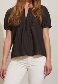 Blusa nera a maniche corte con scollatura a V, caratterizzata da dettagli a righe ricamate e accenti ricamati, abbinata a pantaloni di colore chiaro.