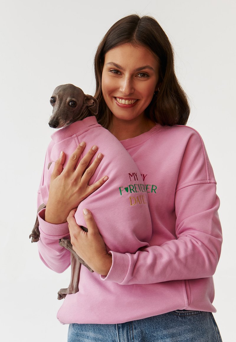 Sweatshirt roz cu text brodat „DATA MEA PENTRU TOTDEAUNA”, purtat de o persoană care ține un câine gri. Materialul pare moale și confortabil.