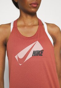 Nike-hihaton toppi ruosteenoranssissa, hihaton malli, jossa suuri vaaleanpunainen ja musta logo rinnassa, pehmeästä, sileäpintaisesta kankaasta valmistettu.