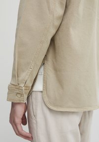 Beige button-up shirt with a relaxed fit, featuring a straight hem, side slits, and a textured fabric. Buttoned cuffs with a single button detail.