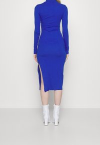 Dlouhý rukáv, bodycon modré šaty s vysokým rozparkem na levé straně, vyrobené z elastického materiálu. Nosené s bílými kotníkovými botami.