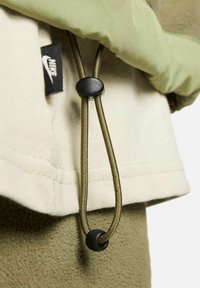 Détail de la veste Nike verte et beige avec un tissu doux, un cordon réglable et une quincaillerie noire sur l'étiquette et le cordon.