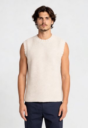 Man met middellang donker haar, gekleed in een mouwloze crèmekleurige gebreide trui en donkere broek, staand tegen een effen lichte achtergrond.