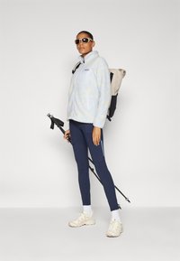 Giacca in pile azzurro chiaro con tasca zip, leggings blu navy con tasche, sneakers beige e occhiali da sole. Porta uno zaino e un bastone da trekking.