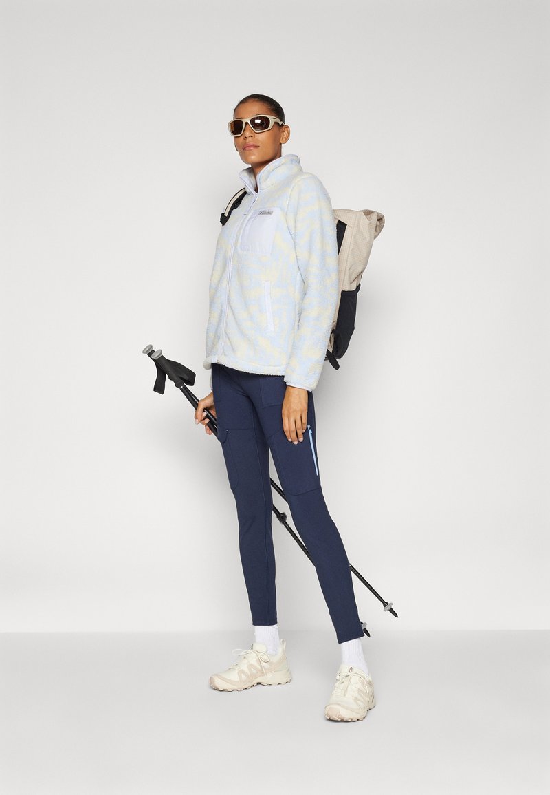 Lichtblauwe fleecejack met een ritszak, marineblauwe leggings met zakken, beige sneakers en een zonnebril. Draagt een rugzak en een wandelstok.