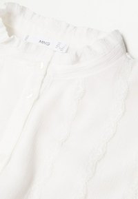 Blouse blanche présentant un col texturé et volé, des rayures verticales subtiles et une fermeture à boutons. Fabriquée en tissu léger et aérien.