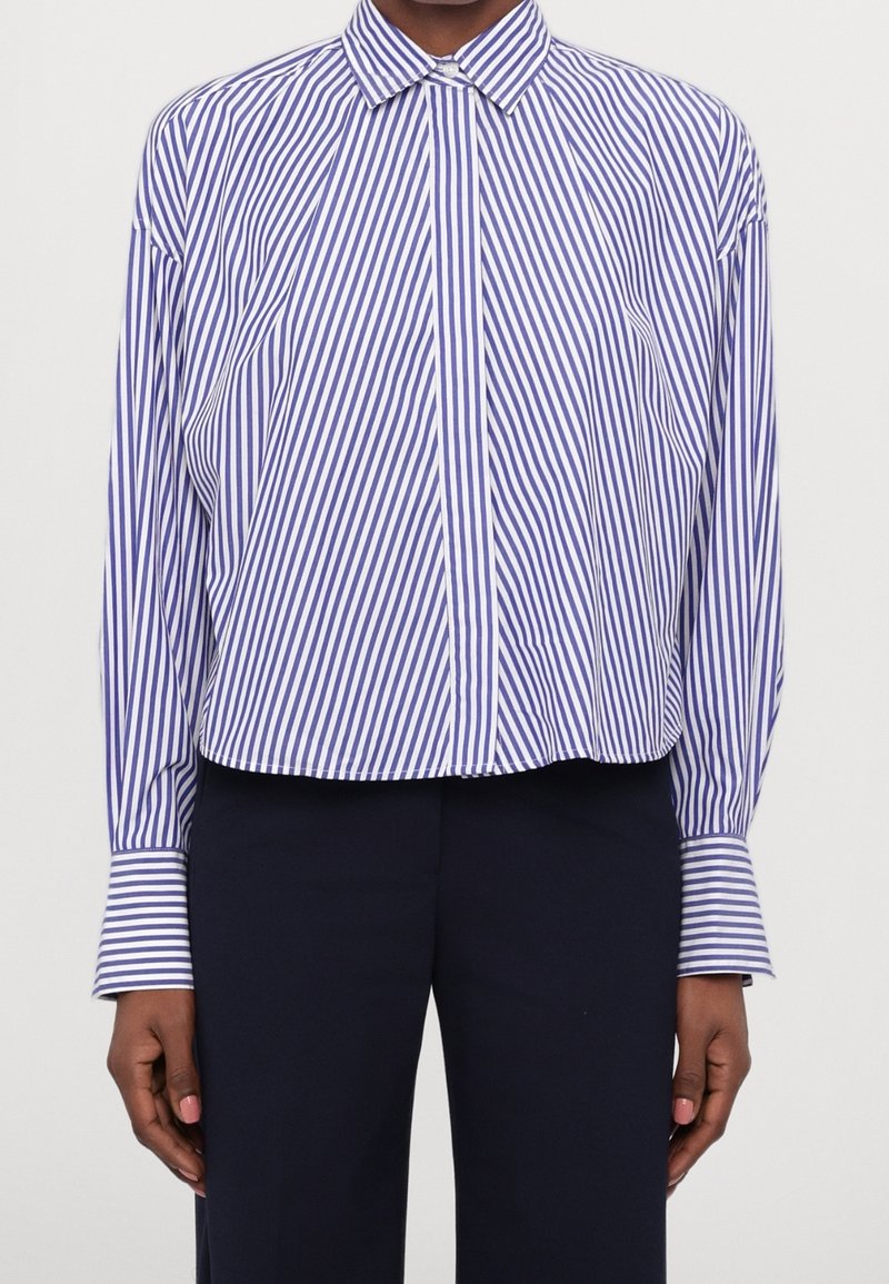 Niebiesko-biała koszula w paski na guziki, z kołnierzykiem, długimi rękawami i krótszym krojem. Tkanina wydaje się gładka i lekka.