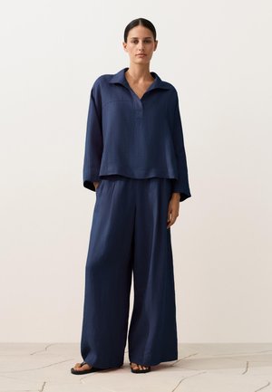Vrouw die een loszittende marineblauwe top met lange mouwen, kraag en wijde broek draagt, staand op een lichte gebarsten vloer tegen een effen achtergrond.