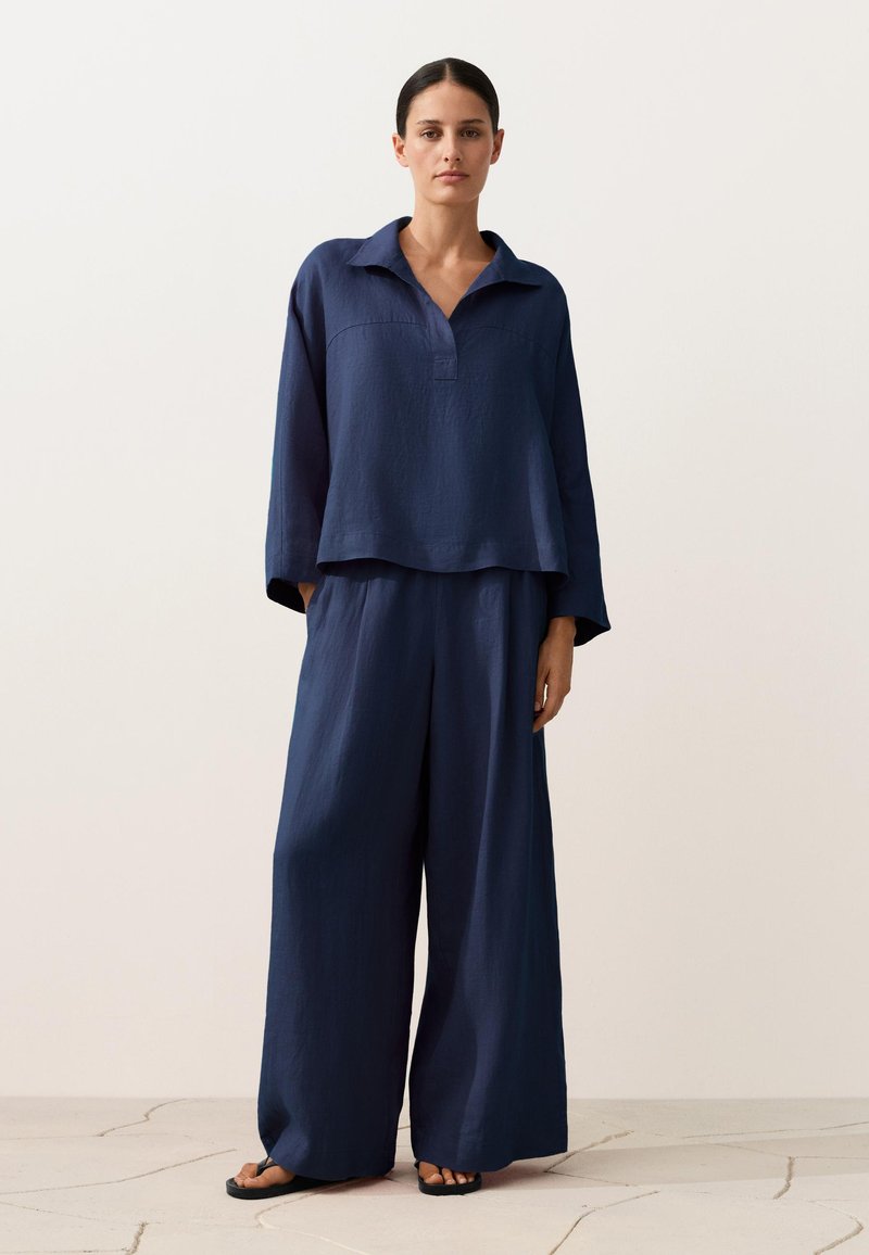 Femme portant un haut ample à manches longues bleu marine avec col et un pantalon à jambes larges, debout sur un sol clair fissuré devant un fond uni.