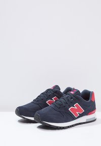 Une paire de baskets New Balance bleu marine avec des logos "N" rouges, des lacets noirs et des semelles blanches sur une surface blanche avec un fond gris clair.