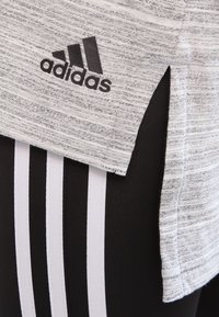 Top sportivo grigio e nero con una finitura texture, con logo Adidas e uno spacco laterale. Abbinato a leggings neri con strisce bianche.