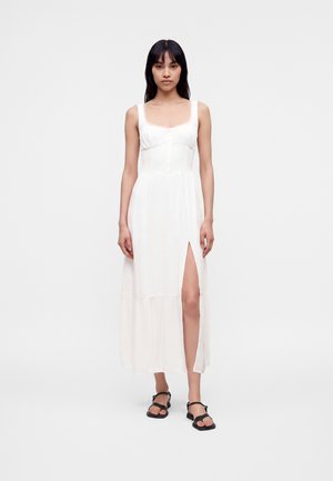 Frau mit langen dunklen Haaren, die ein ärmelloses weißes Kleid mit Knöpfen und einem vorderen Schlitz trägt, kombiniert mit schwarzen flachen Sandalen, vor weißem Hintergrund stehend.