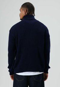 Donkerblauwe, ribgebreide trui met een hoge kraag, losse pasvorm en lange mouwen, gedragen over een wit shirt, met een textuurrijke stof.