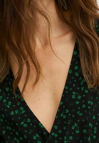 Robe noire avec un décolleté en V et un motif floral vert, en tissu lisse et avec une coupe fluide, mettant en valeur le décolleté et l'épaule.