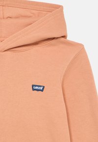 Φούτερ με κουκούλα σε ροδακινί απόχρωση, κατασκευασμένο από απαλό ύφασμα, με μπλε λογότυπο Levi's στο μπροστινό μέρος. Ο σχεδιασμός περιλαμβάνει κουκούλα με κορδόνι και ριμπ λεπτομέρεια.