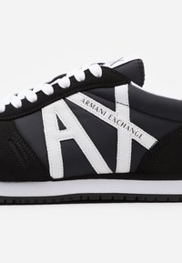 Sorte og hvite joggesko med overdel i semsket skinn og nylon, fremtredende "AX" logo, og hvite snøringer på en gummisåle med mønster for godt feste.