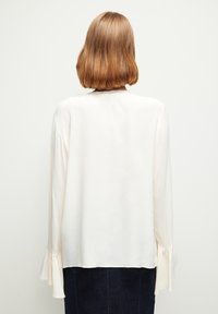 Frau mit schulterlangen, hellbraunen Haaren, die eine lockere weiße Bluse mit langen Ärmeln und Rüschenbündchen sowie einen dunklen Rock trägt, sie schaut weg.