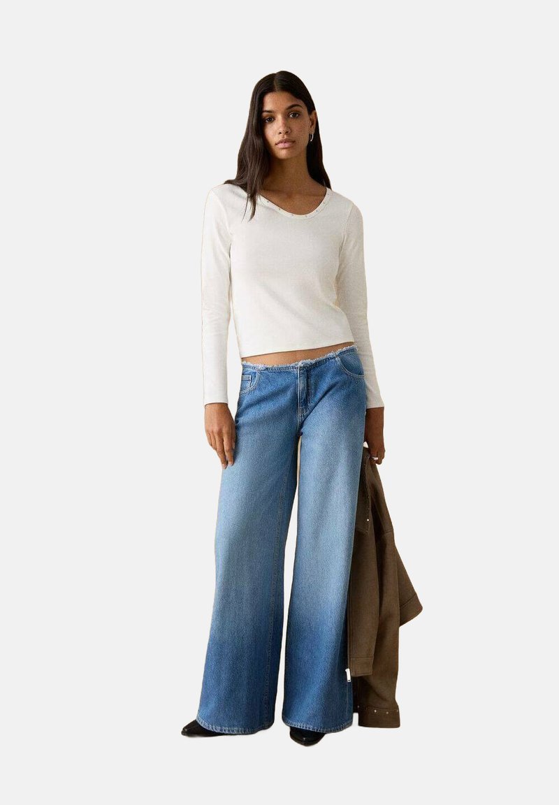 Top bianco a maniche lunghe abbinato a jeans larghi a gamba larga e slavati in blu. La modella tiene in mano una giacca marrone. Gli articoli sono casual e di stile contemporaneo.