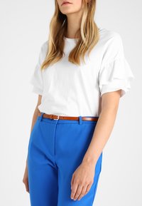T-shirt en coton blanc avec des manches courtes froncées, associé à un pantalon bleu vif et une ceinture en cuir marron avec une boucle en argent.