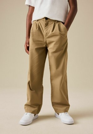 Osoba ubrana w beżowe spodnie o szerokich nogawkach, białe trampki i białą koszulę, stojąca z jedną ręką w kieszeni na jednolitym tle.