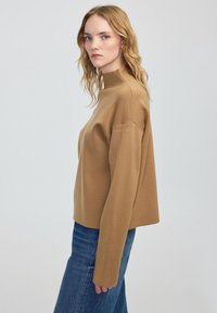 Světle hnědý rolák se uvolněným střihem, vyrobený z měkké tkaniny. Má dlouhé rukávy a zkrácený lem, kombinovaný s modrými džíny.