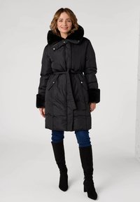 Manteau d'hiver noir matelassé avec un col en fourrure, taille nouée, poches zippées et poignets en fausse fourrure noire. Porté avec un jean foncé et des bottes hautes.
