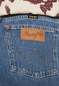 Blå denimjeans med en brun läderlapp märkt "Wrangler", med detaljer på bakfickorna och guldfärgade sömmar.