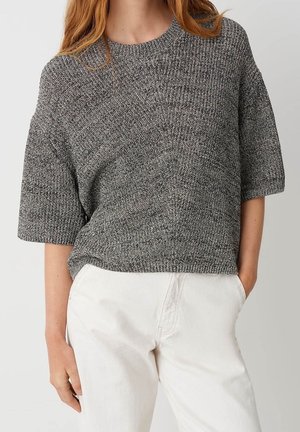 Femme portant un pull en maille gris à manches courtes et un pantalon blanc taille haute, debout avec une main dans la poche.