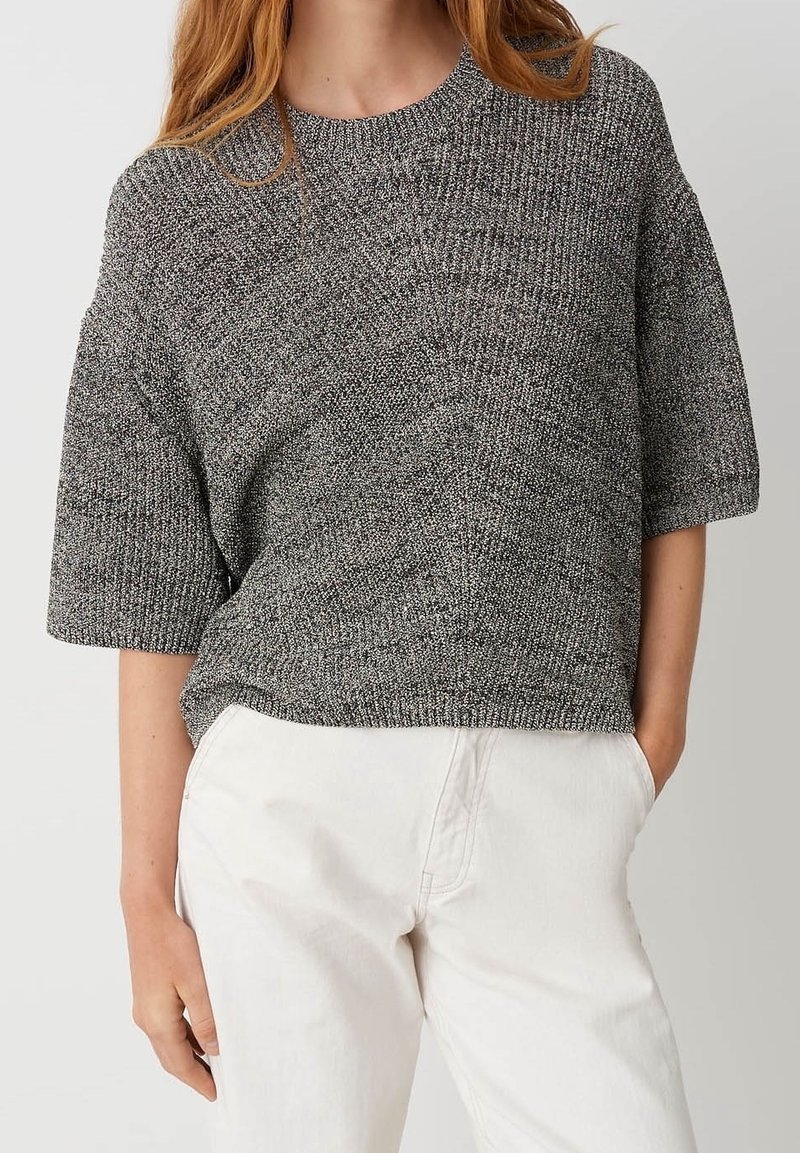 Femme portant un pull en maille gris à manches courtes et un pantalon blanc taille haute, debout avec une main dans la poche.