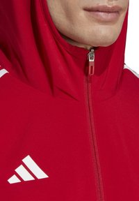 Rode zip-up hoodie van zachte stof, met een hoge kraag en witte strepenhals accenten op de schouders. Een opvallend wit logo is zichtbaar.