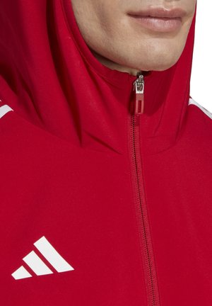Rode zip-up hoodie van zachte stof, met een hoge kraag en witte strepenhals accenten op de schouders. Een opvallend wit logo is zichtbaar.