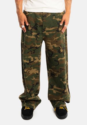 Homme portant un pantalon de camouflage ample vert et marron, avec des chaussures noires et des lacets jaunes sur fond blanc.