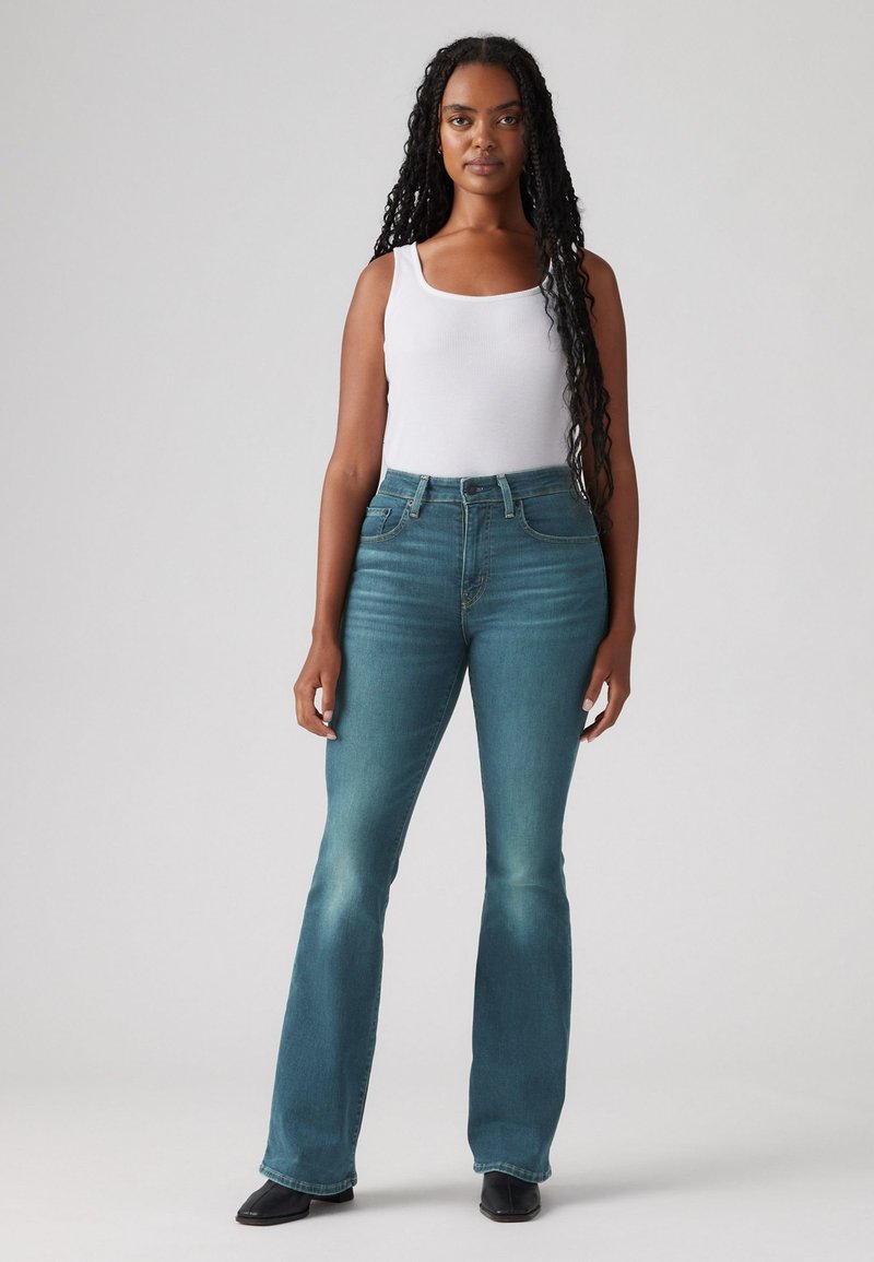 Jeans in denim a vita alta e svasati in blu sbiadito con un leggero gradiente. Presentano un classico design a cinque tasche e passanti per cintura. Abbinati a un top bianco senza maniche.