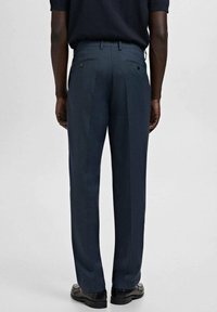Hombre con pantalones de vestir azul marino y zapatos negros, de pie contra un fondo gris liso, visto desde atrás.
