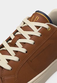 Baskets en cuir marron avec lacets crème, œillets dorés et semelle blanche, présentant des détails cousus et une étiquette de languette en cuir estampillée de la marque.