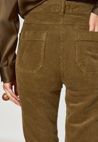 Pantaloni in velluto a coste marroni con righe verticali, due tasche posteriori e un alto punto vita. Mano appoggiata sul fianco, che mette in mostra un anello.