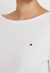 Nahaufnahme einer Person, die ein weißes Hemd mit einem kleinen rot-, blau- und weißfarbenen rechteckigen Logo auf der Brust trägt.