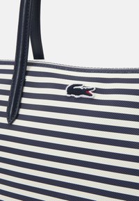 Sac fourre-tout rayé en matériau synthétique bleu marine et crème, avec un logo Lacoste. Le design comprend des poignées en cuir robustes.