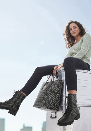 Black ankle boots with patterned fabric sides, round toe, and laces. Large black tote with geometric design. Model in a green ribbed top and dark pants.