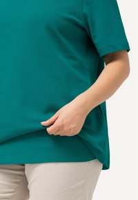 Teal kort mouwen top van zachte stof, met een relaxte pasvorm en zijsplit. Hand houdt de zoom vast, terwijl lichtbeige broek gedeeltelijk zichtbaar is.